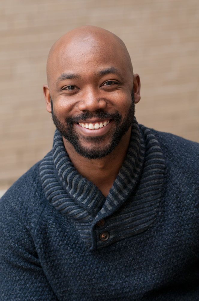 A smiling bald man with a beard wearing a navy sweater poses for a warm professional headshot.