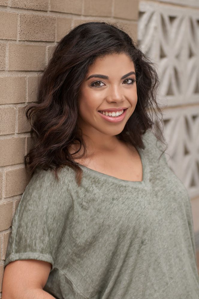 A smiling young woman with wavy dark hair wearing a sage green top poses near a brick wall.