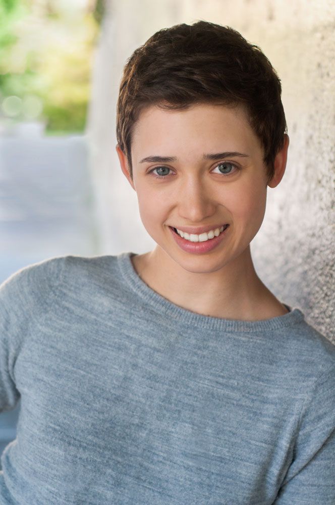 Smiling young woman with short dark hair wearing a gray sweater leaning against a light gray wall.