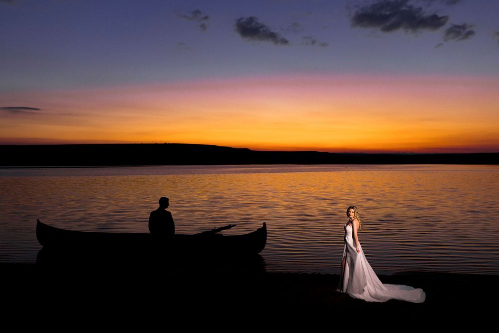 Bride and groom silhouetted at sunset by a lake with canoes, golden sky reflecting on calm water.