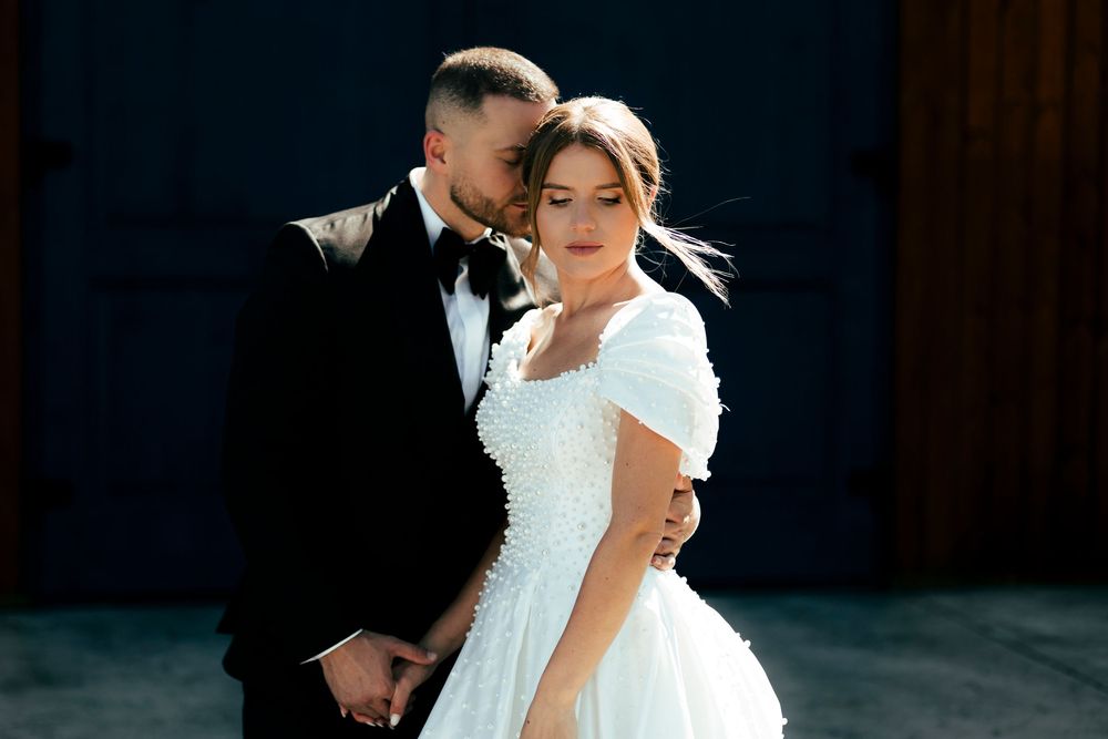 Elegant bride in white lace ball gown and groom in black tuxedo embracing tenderly outdoors.
