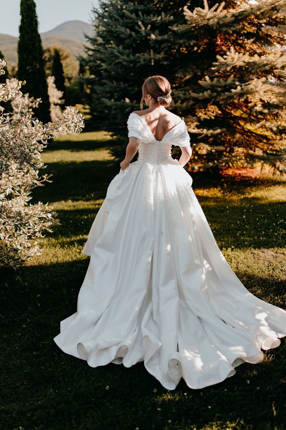 Bride in elegant white off-shoulder ball gown with long train stands in lush garden with mountain views.