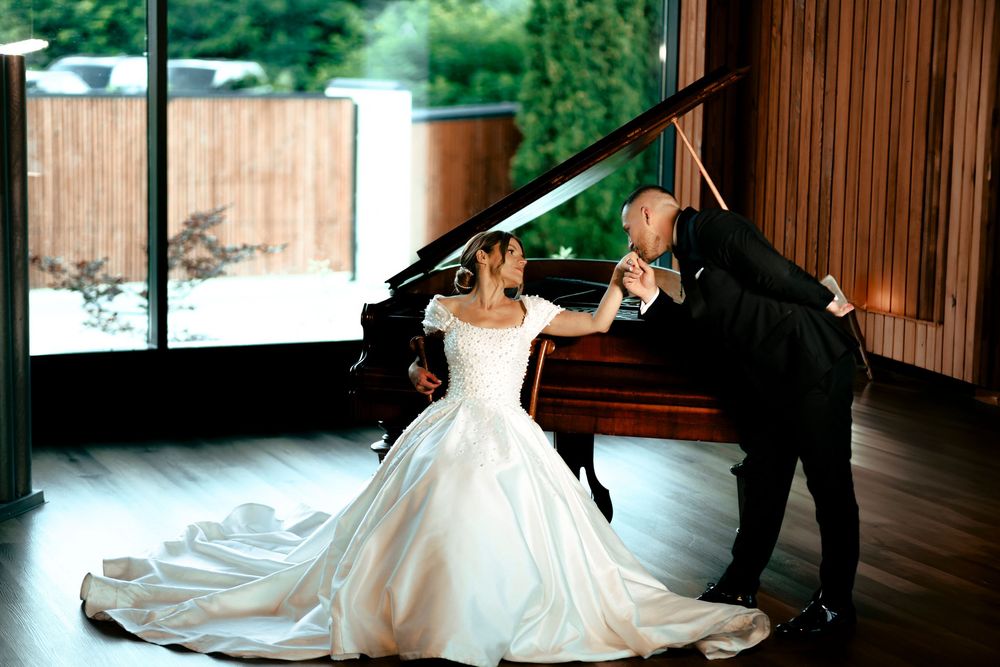 Bride in elegant ball gown and groom in tuxedo share romantic moment at grand piano in modern venue.