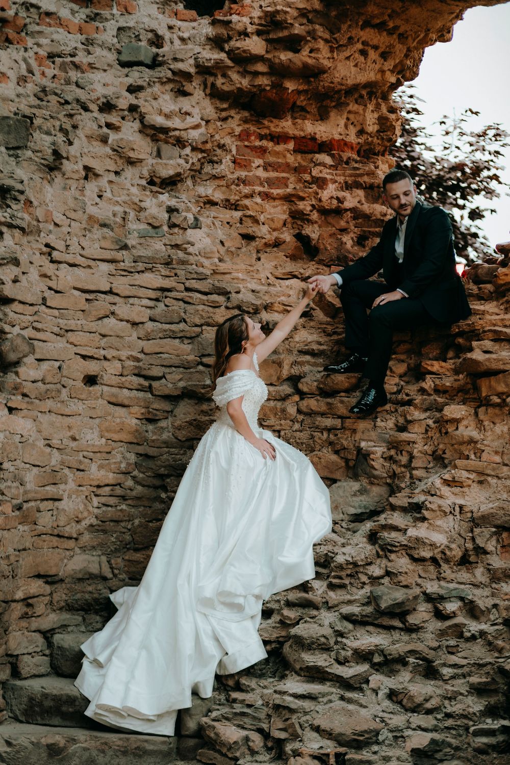 Bride in white gown reaches up to groom sitting on ancient stone ruins wall in romantic wedding photo.