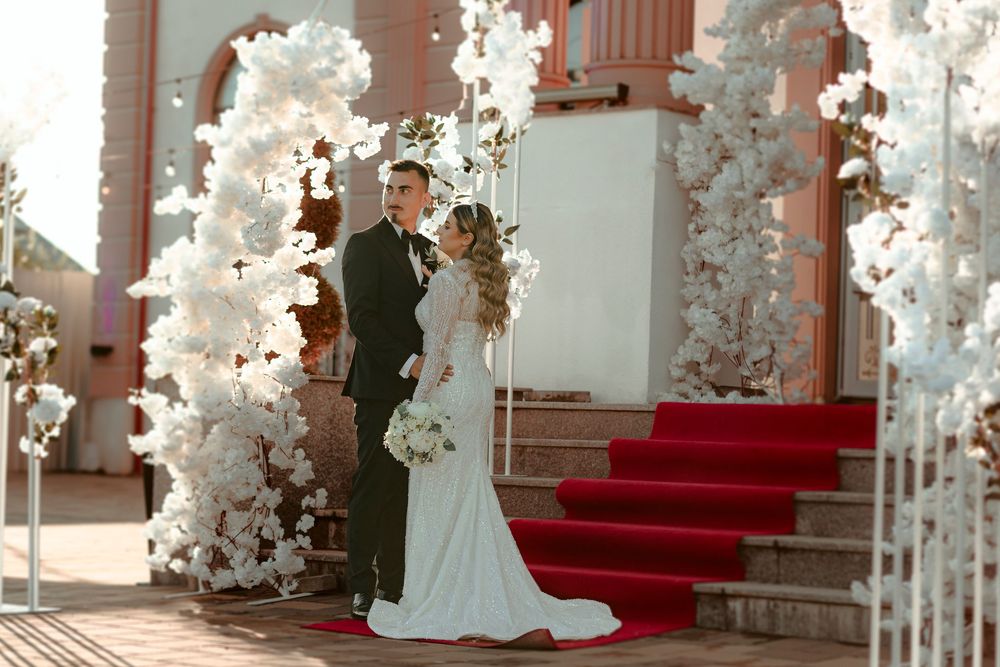 Bride and groom pose under white floral arch with red carpet steps at elegant outdoor wedding venue.