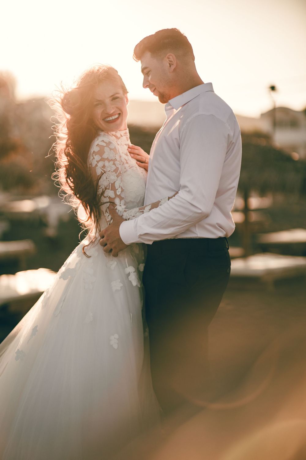 A joyful bride in a white lace gown laughs with her groom at golden hour, embracing outdoors.