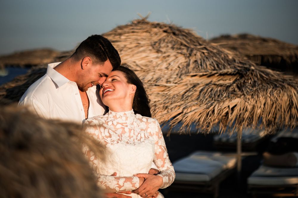 Romantic couple embracing at sunset near thatched umbrella on a beach, sharing a tender moment in white outfits.