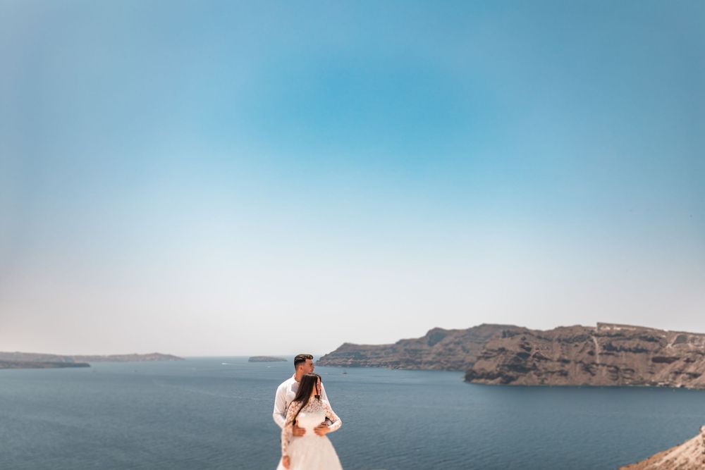 Couple in wedding attire standing on a cliff overlooking the stunning blue waters and volcanic landscape of Santorini, Greece.