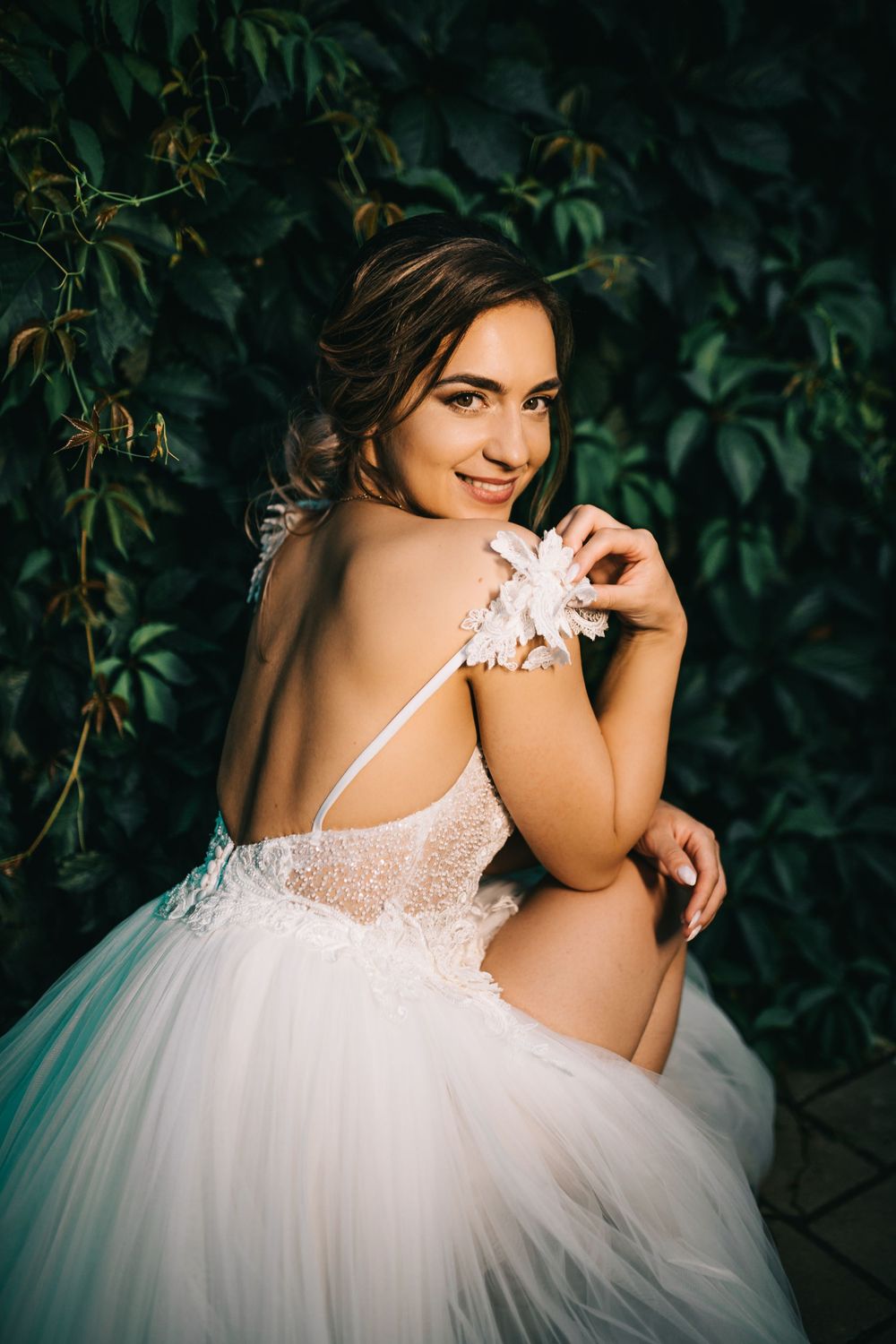 Smiling bride in white lace tulle gown with floral detail poses against lush green ivy wall backdrop.
