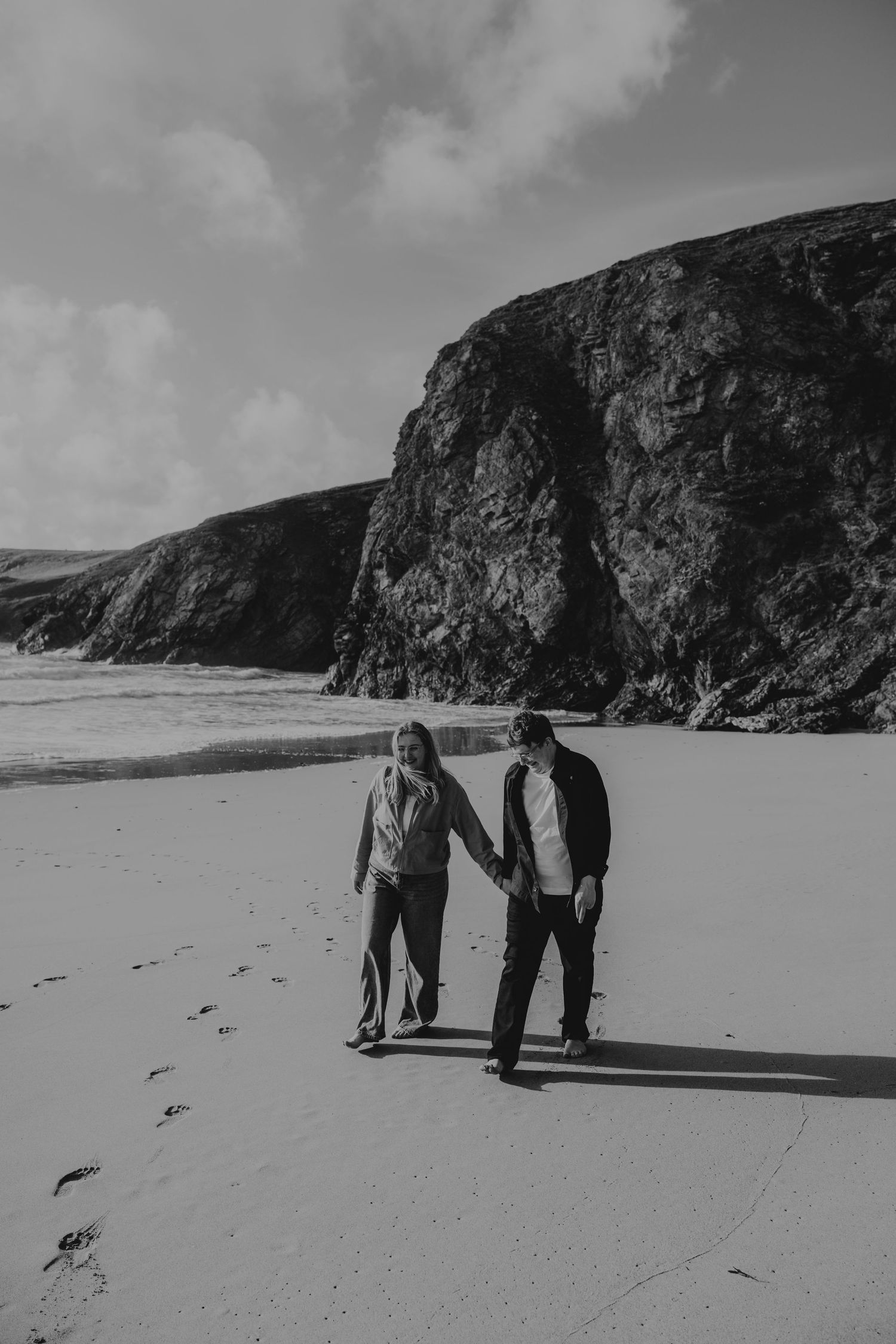 Black and white photo of a couple holding hands while walking on a sandy beach with dramatic rocky cliffs in the background.