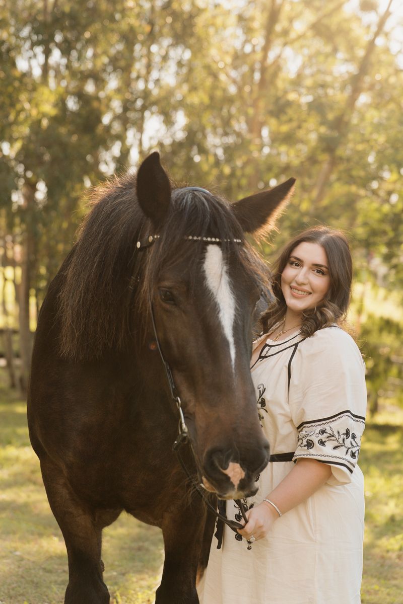 Young woman in cream dress smiling beside a dark brown horse outdoors in golden sunlight during a photoshoot with a horse in sydney