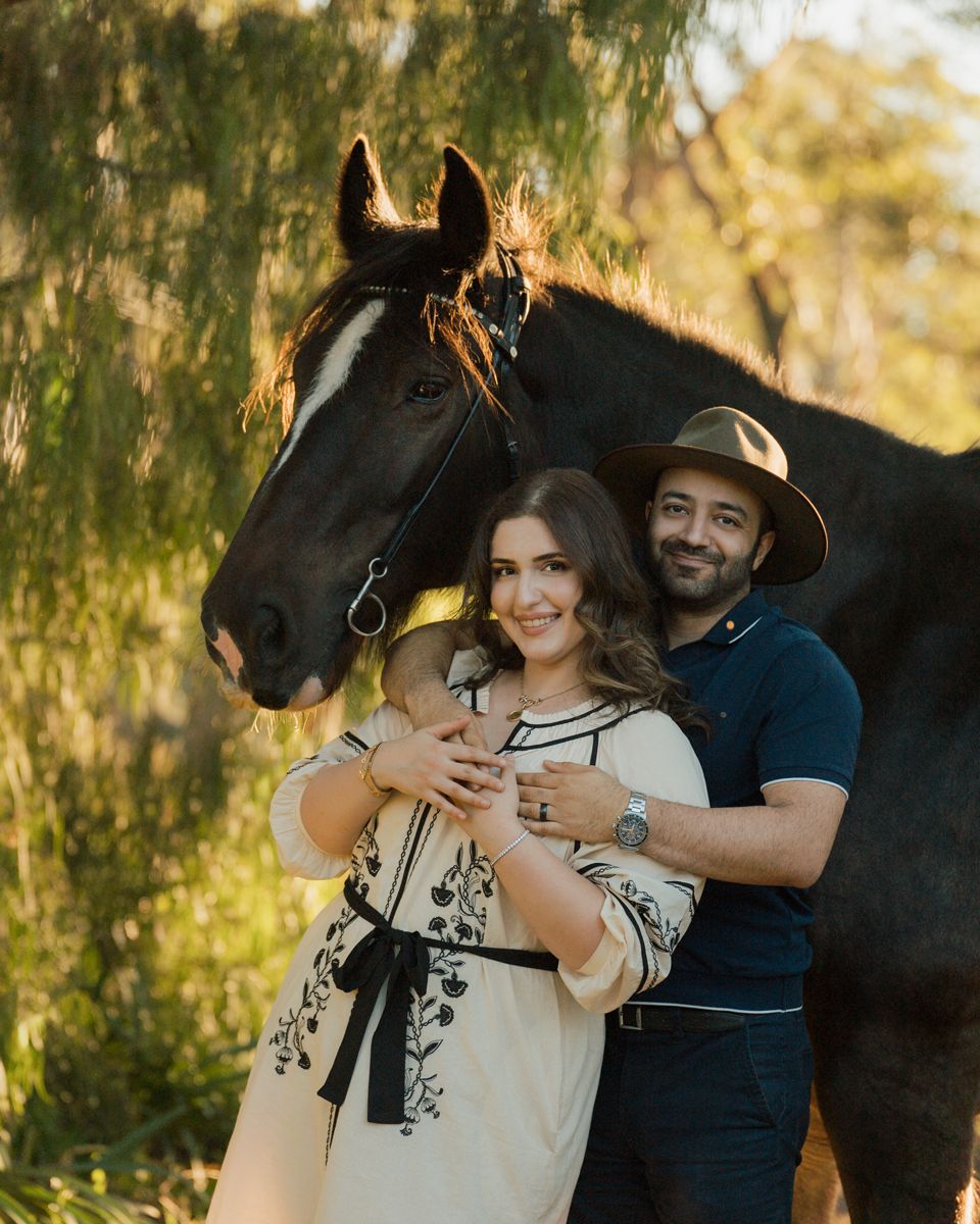 Couple smiling together beside a horse outdoors in golden sunlight, woman in floral dress, man in navy shirt. photoshoot in sydney