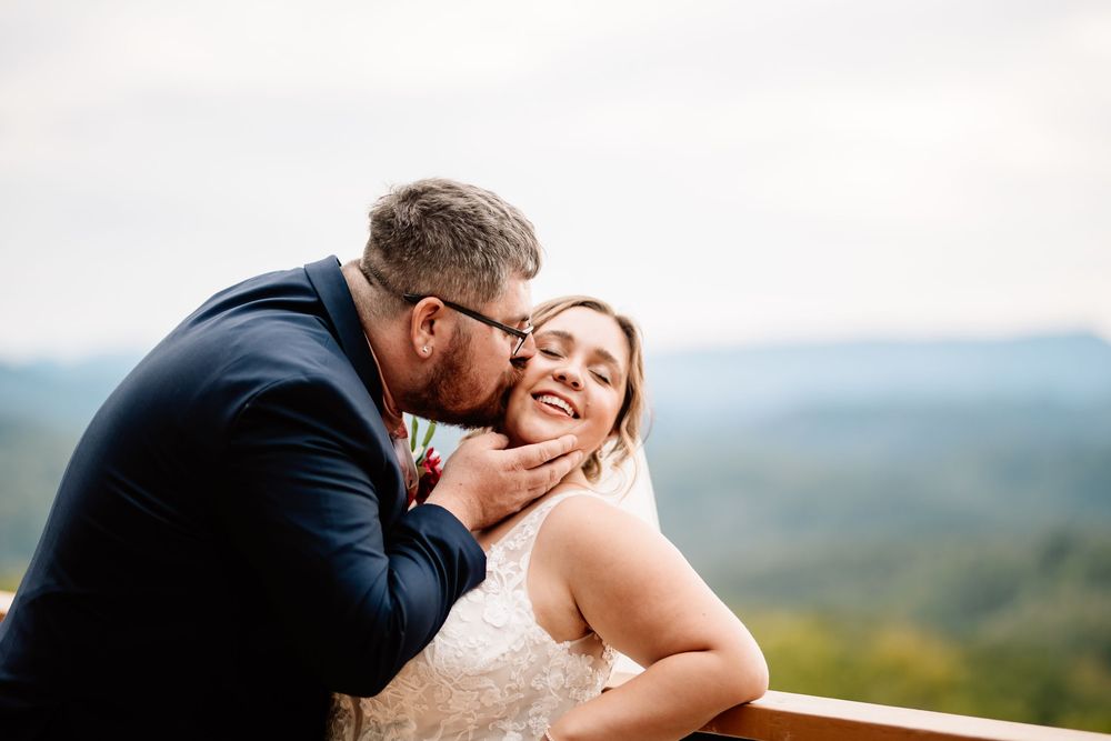 Groom tenderly kisses bride on cheek as she smiles with lush mountain landscape behind them.