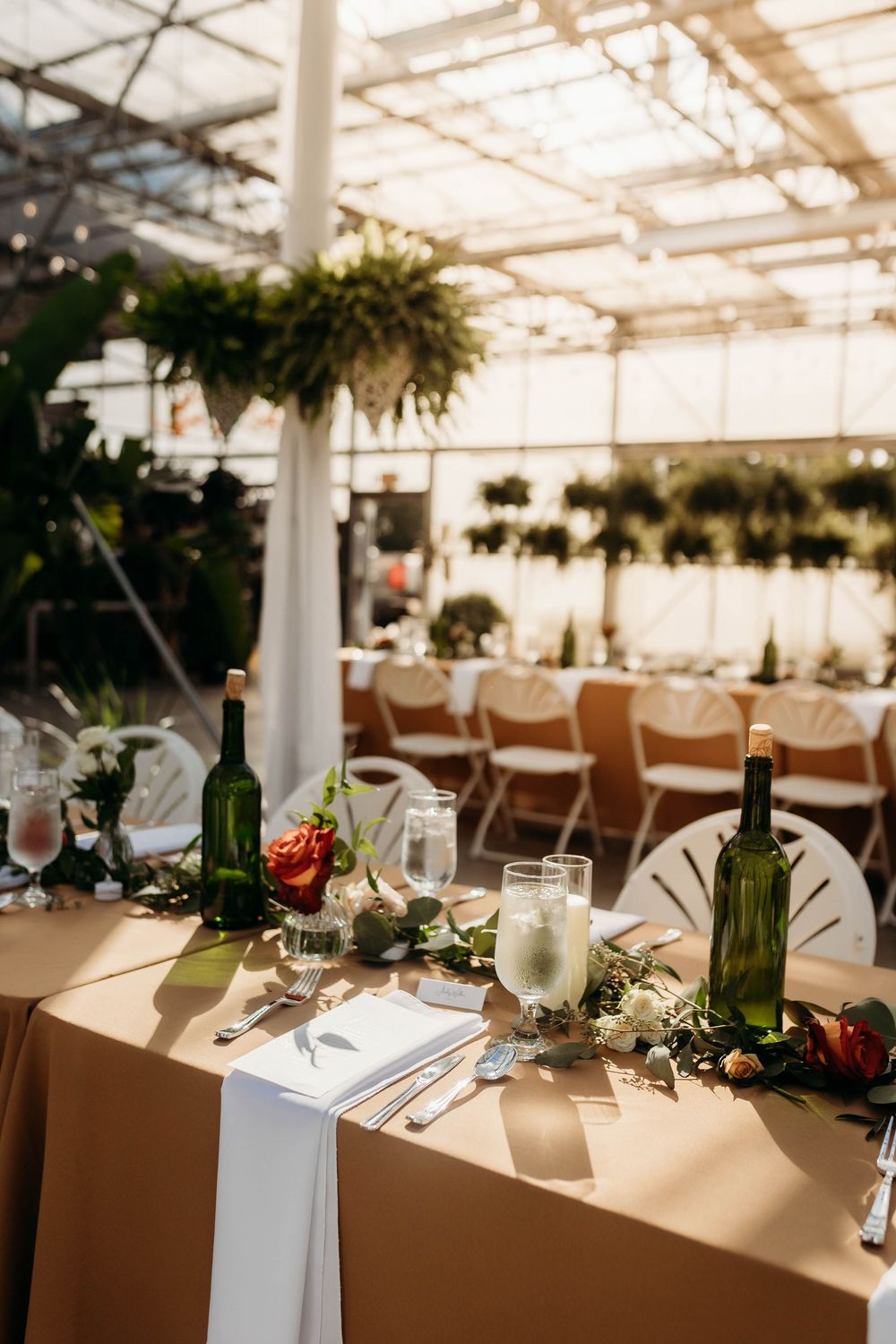 Elegant greenhouse wedding reception table with green wine bottles, floral garland, candles, and folding chairs in warm sunlight.
