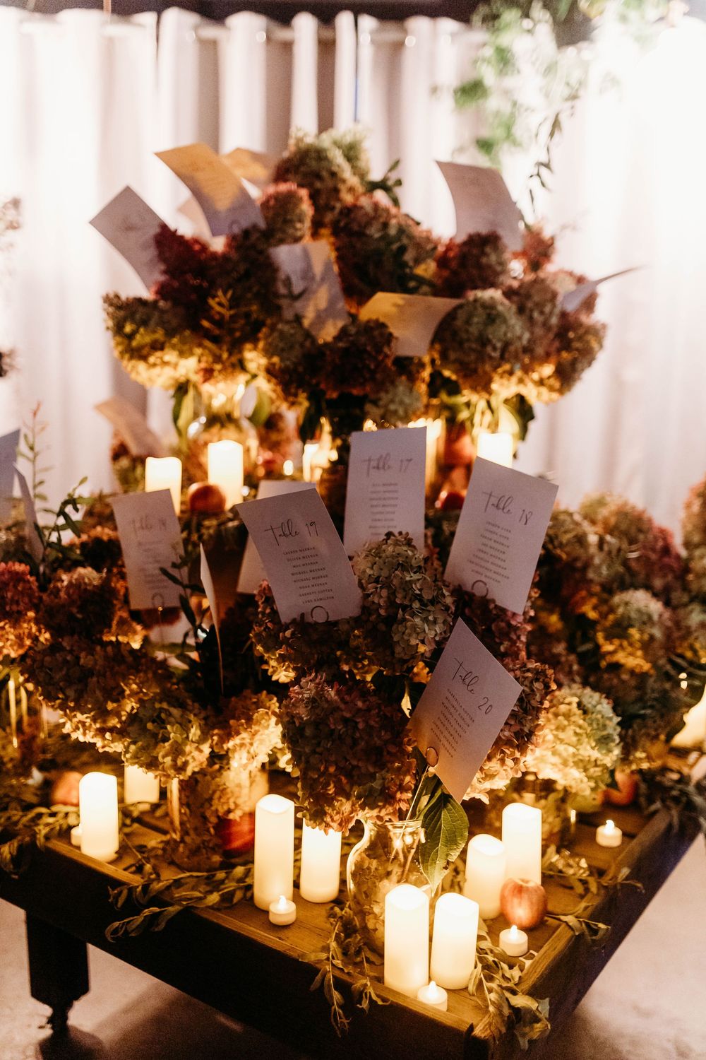 Elegant wedding seating chart display with candles and dark floral hydrangea arrangements in warm amber lighting.
