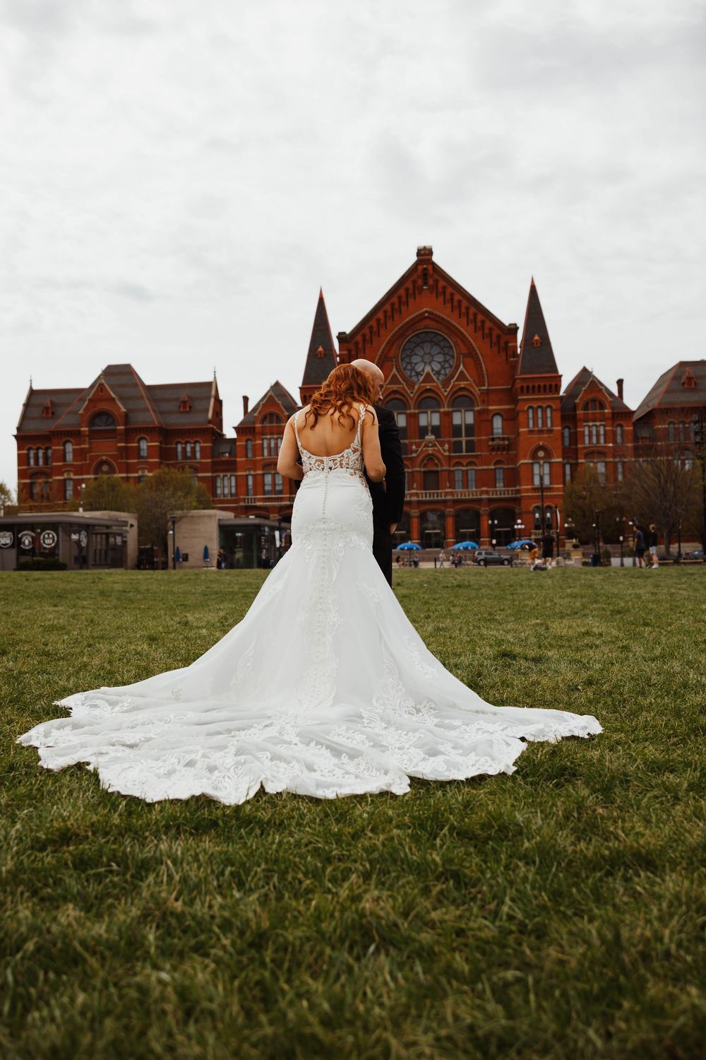 Bride in white lace mermaid gown with long train stands with groom before historic red brick building.
