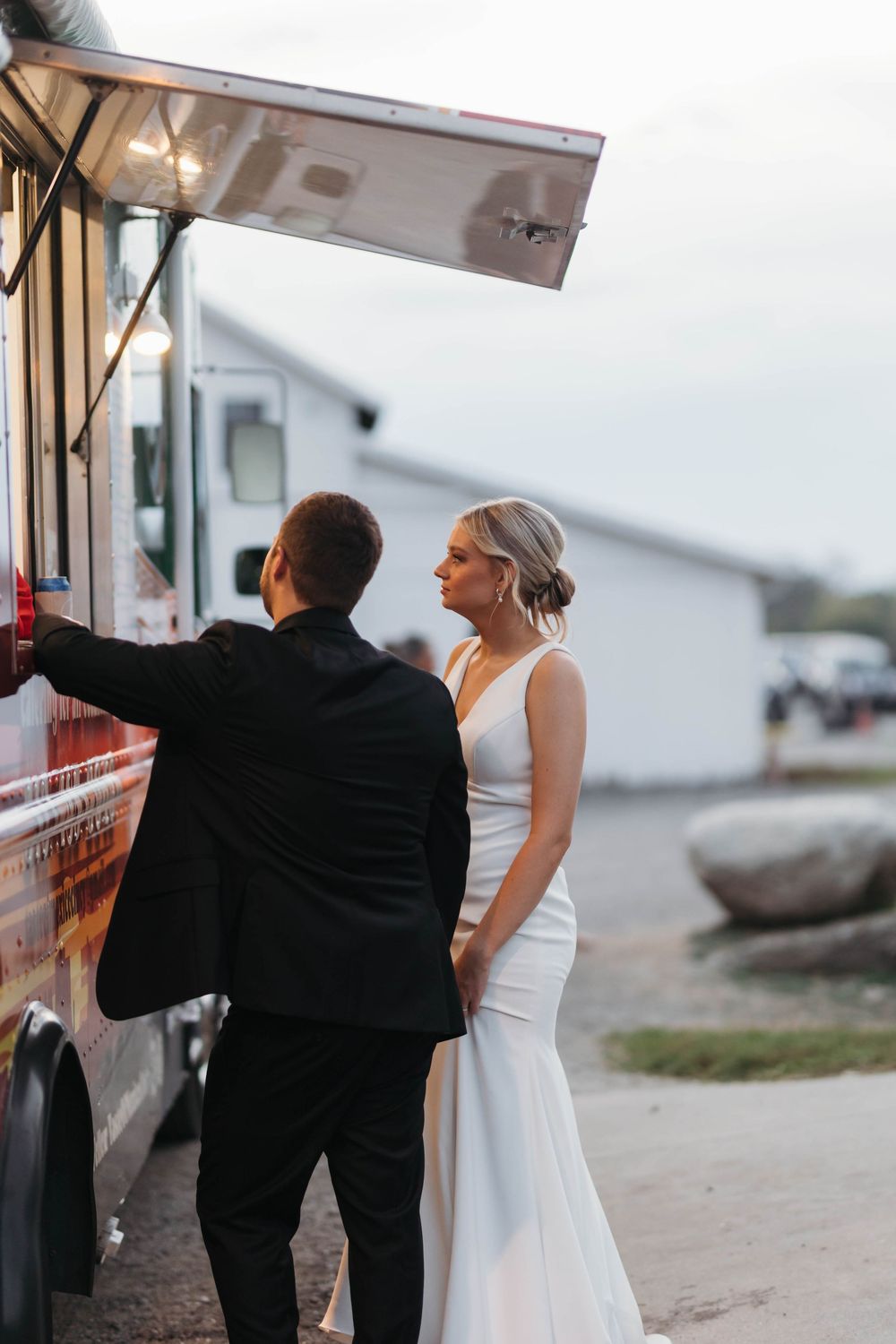Bride in sleek white gown and groom in black suit ordering from a food truck at their outdoor wedding reception.