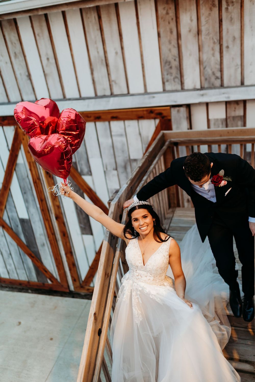 Smiling bride in a lace gown holds red heart balloons on stairs as groom in black suit leans in beside her.