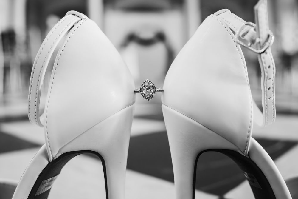 Black and white photo of white bridal heels with a diamond engagement ring placed between them.