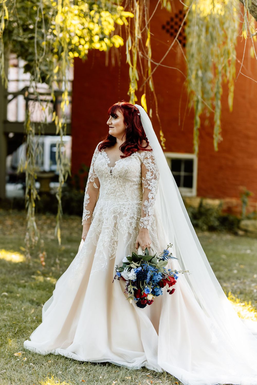 Bride in lace long-sleeve wedding gown holding blue and red bouquet under weeping willow tree.