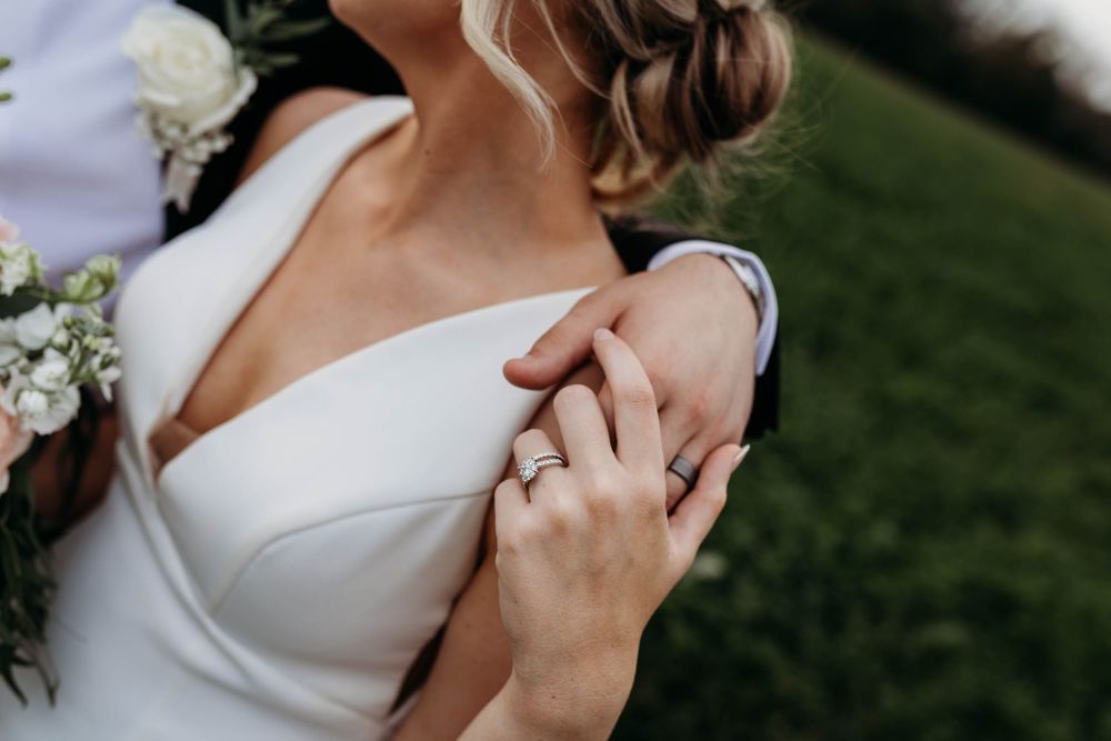 Close-up of bride in white off-shoulder gown with diamond ring, holding groom's hand at outdoor wedding.