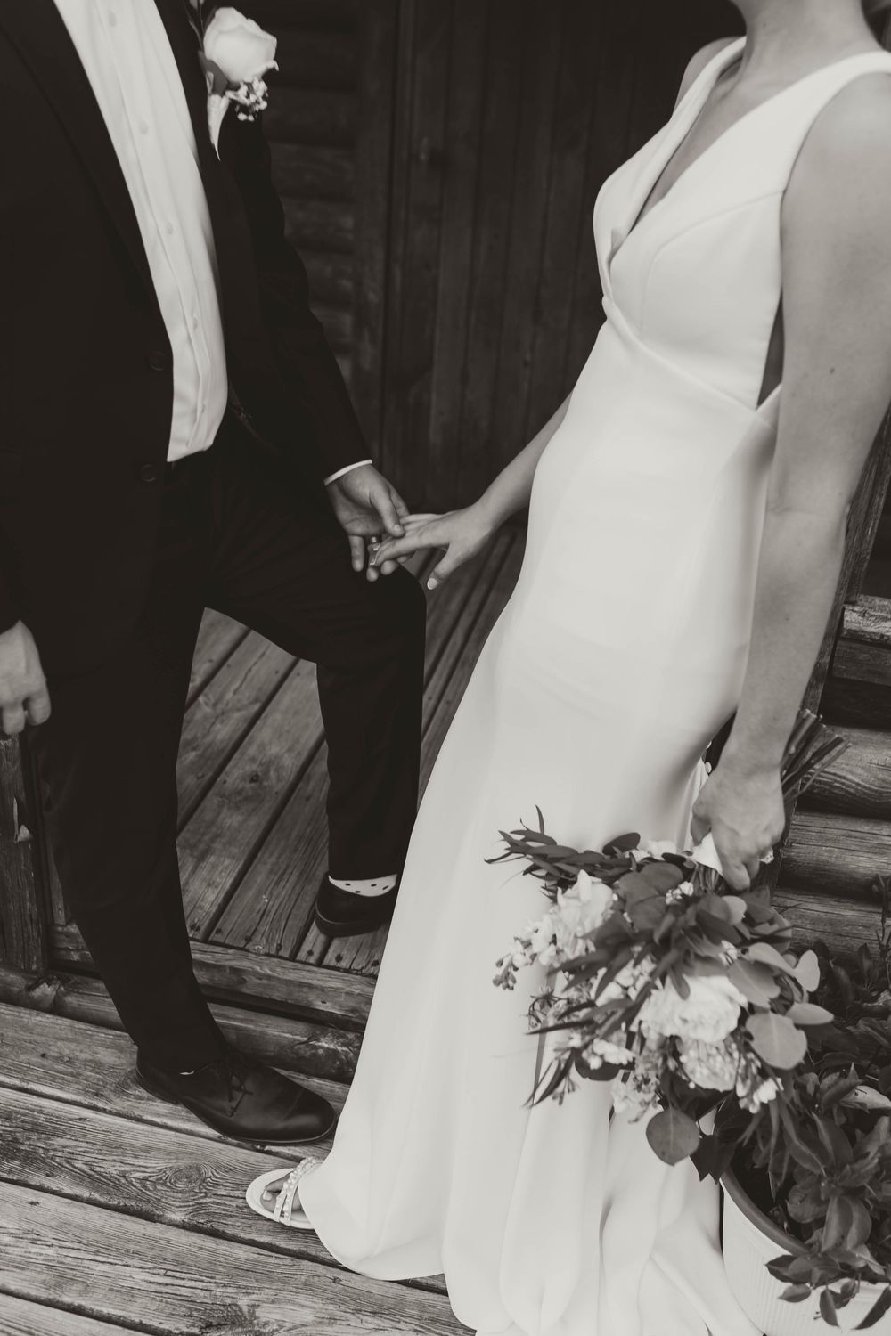 Black and white photo of bride and groom holding hands, bride in elegant gown holding floral bouquet on wooden deck.