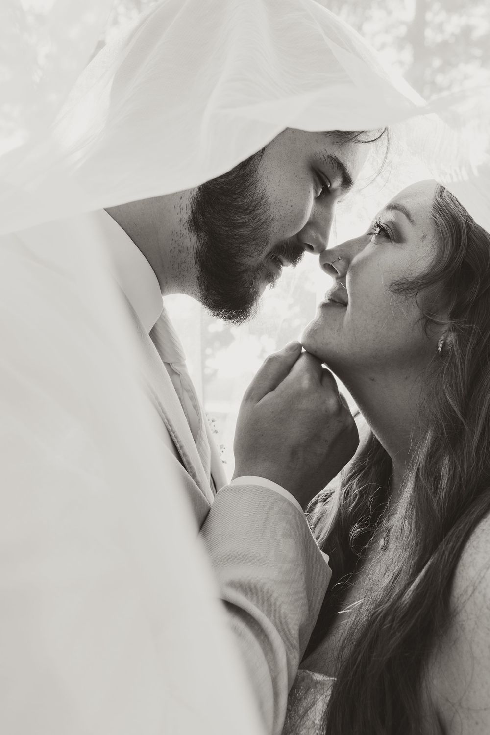 Black and white photo of a romantic couple almost kissing under a bridal veil on their wedding day.