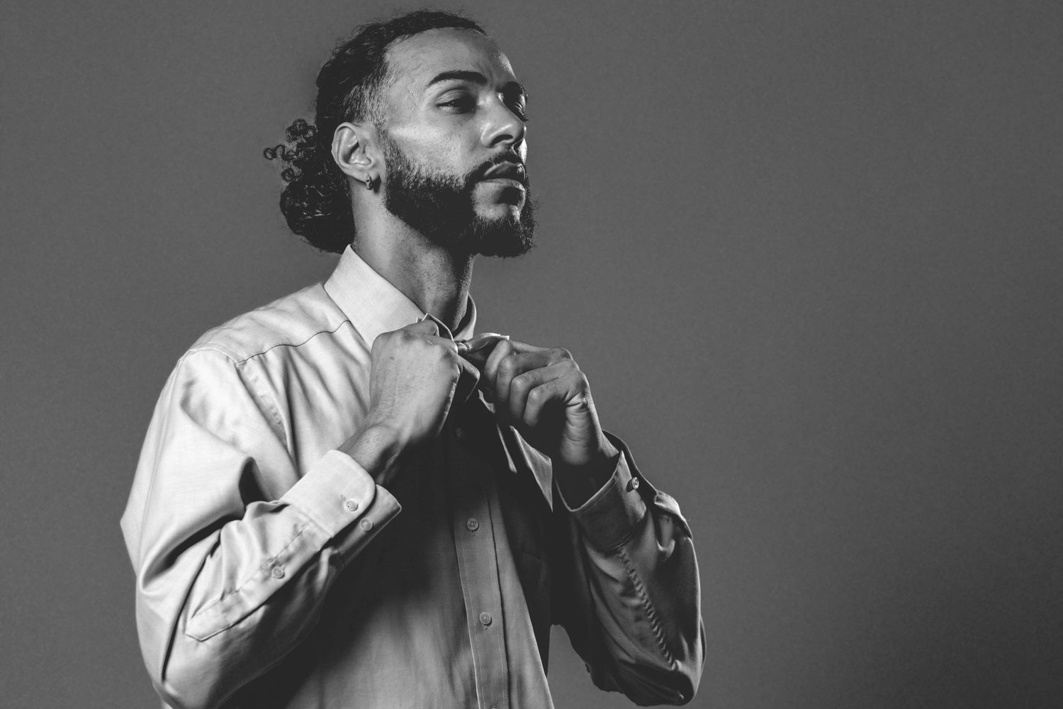 Black and white photo of a young man adjusting his collar, wearing a button-up shirt against a gray background.