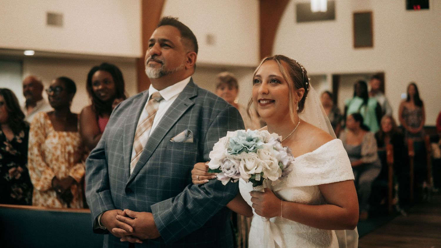 Bride walking down the church aisle smiling with bouquet accompanied by suited gentleman.