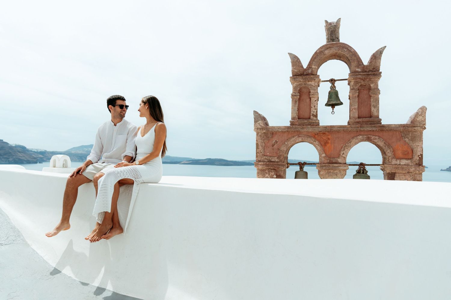 Couple in white outfits sitting on a white wall by a rustic bell tower in Santorini, Greece.