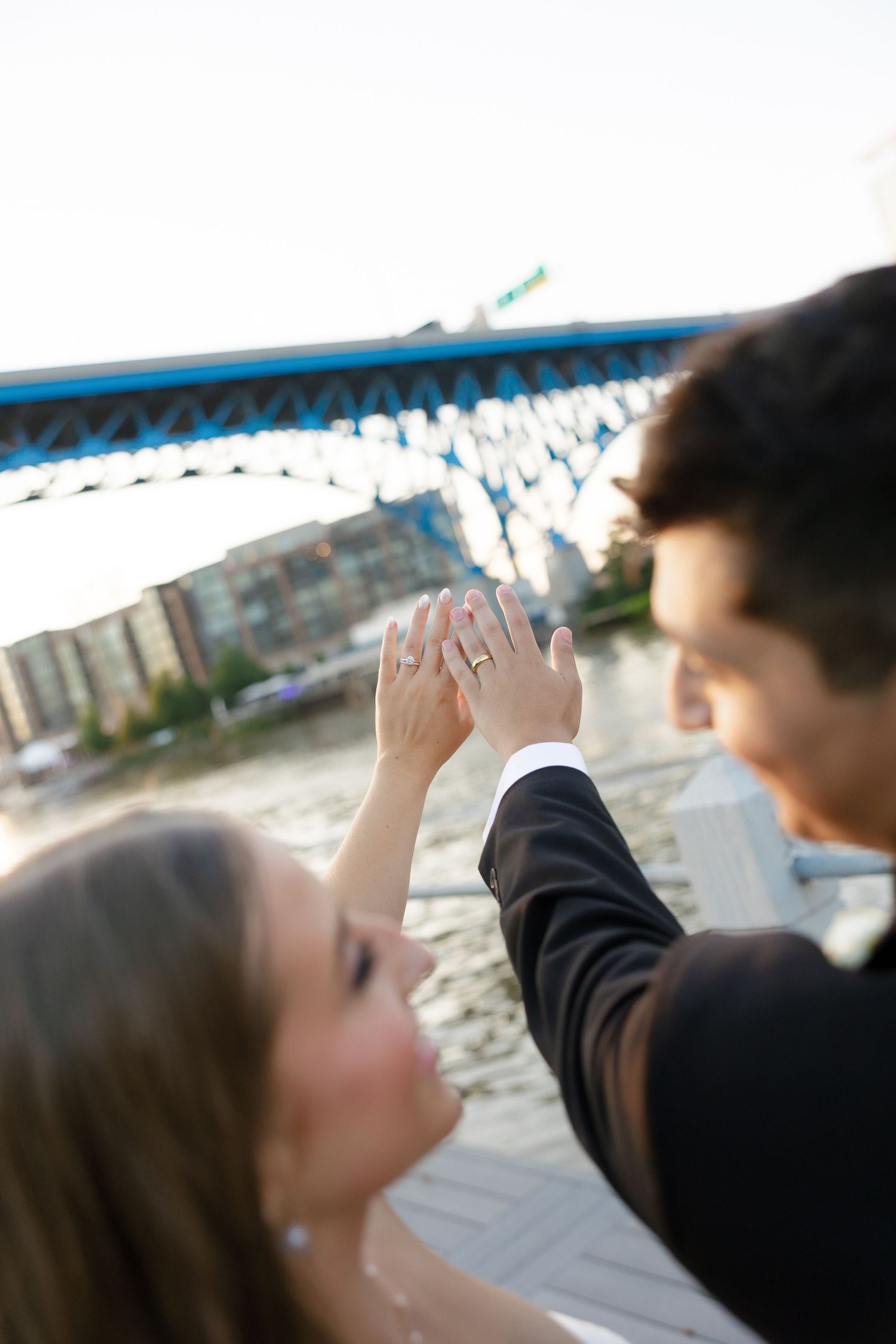 Bride and groom holding hands near a blue bridge waterfront, showcasing wedding ring during romantic outdoor photo session.