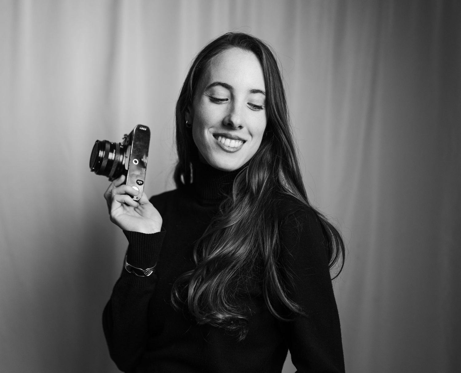 Black and white portrait of a smiling woman holding a camera, wearing a black turtleneck against a light backdrop.