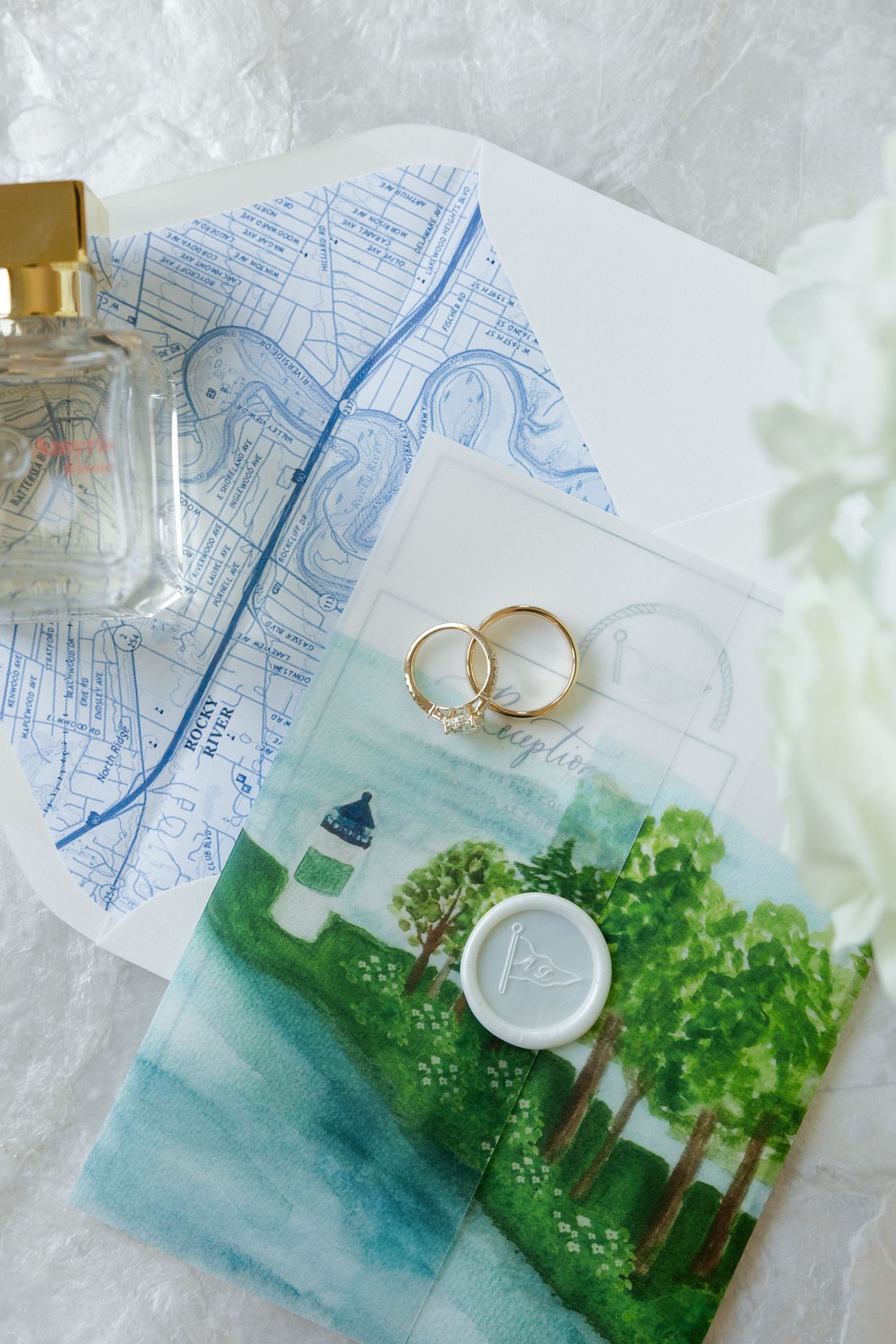 Wedding flat lay featuring gold rings, watercolor invitation, wax seal, and perfume bottle.