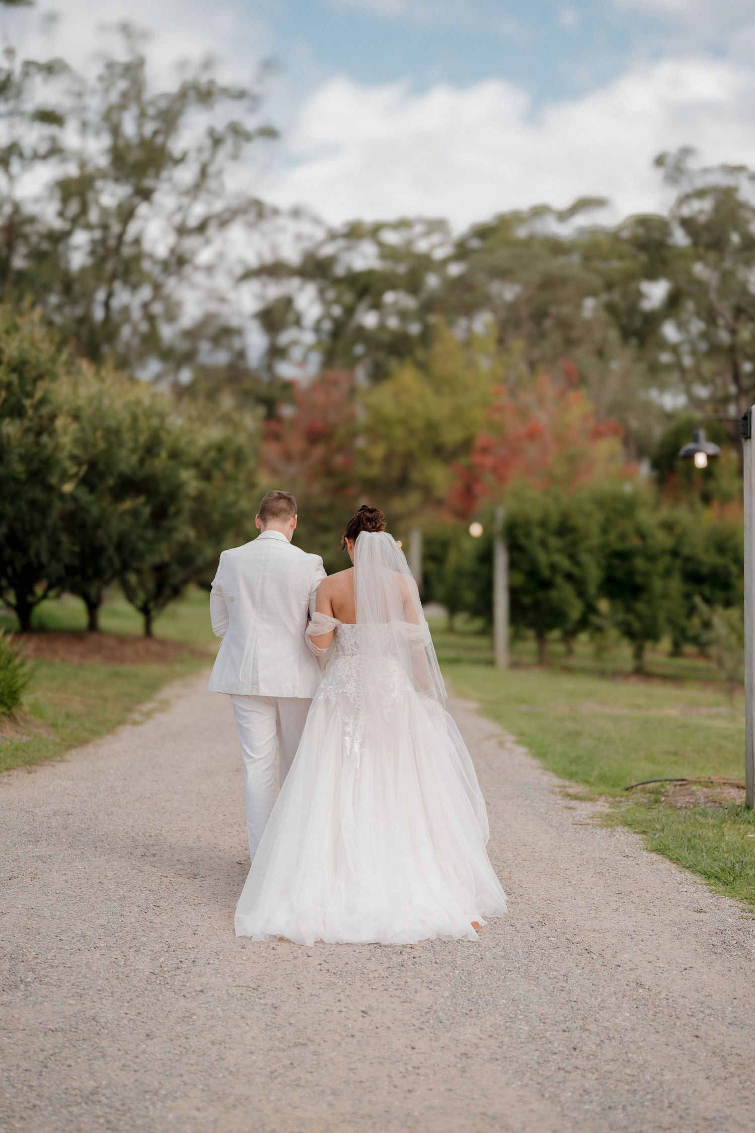 A bride and groom walk hand in hand down a tree-lined path, viewed from behind, on their wedding day.
