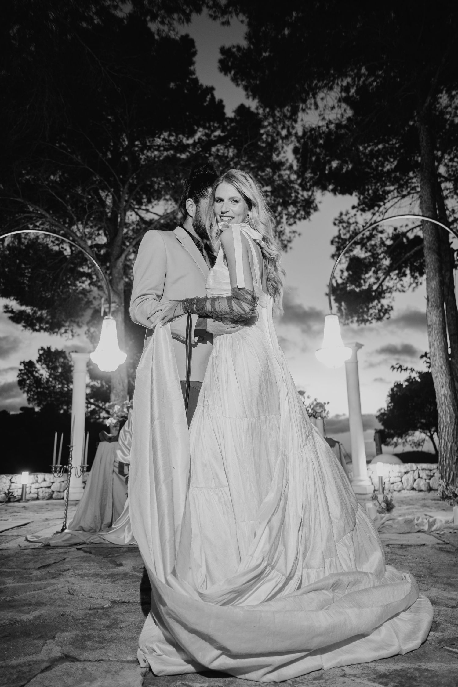Black and white wedding photo of couple embracing outdoors at dusk with illuminated candles and trees in background.