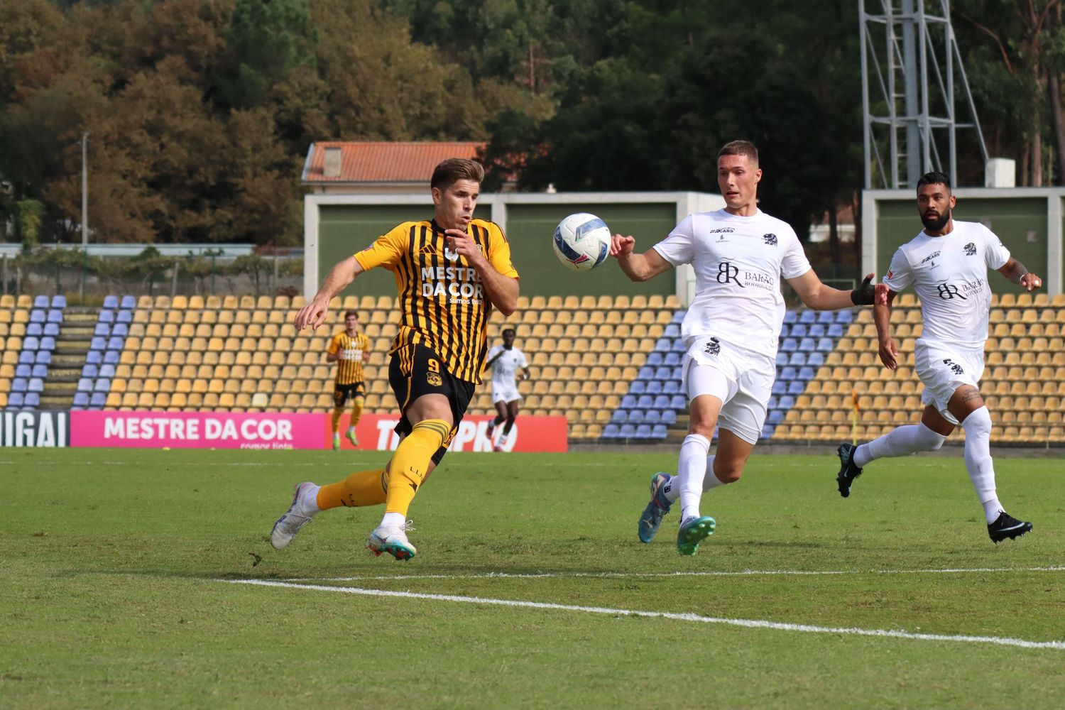 Soccer players compete for the ball during a match on a green field with stadium seating visible.