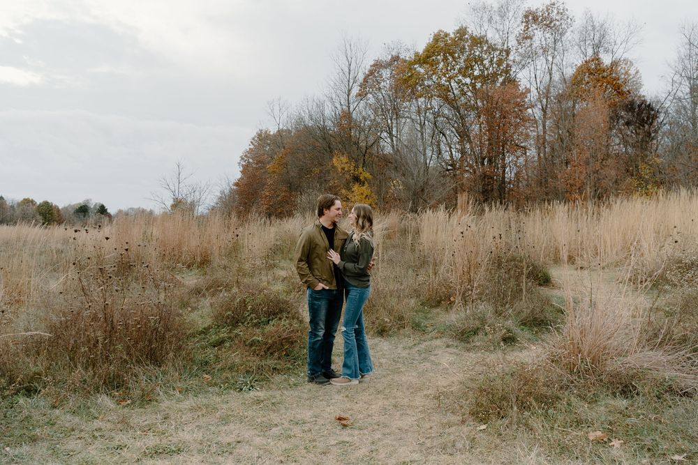 Romantic couple sharing tender moments in a field of tall autumn grass with fall foliage in background.