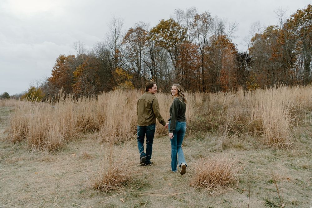 Two people walk together through a golden autumn field with tall dried grass and trees in the background.