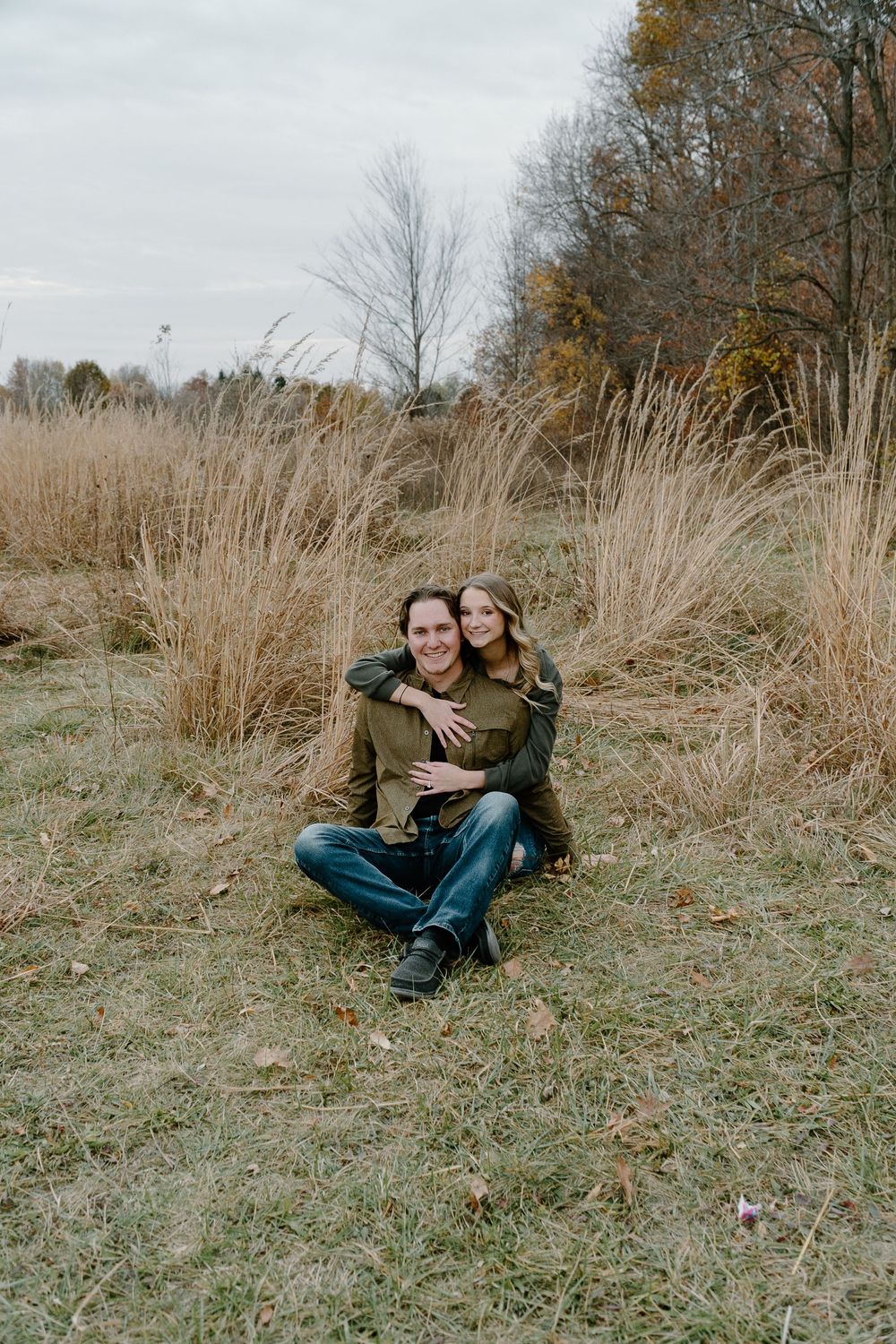 Two people in casual attire sitting together on a grassy hillside during autumn with bare trees in the background.