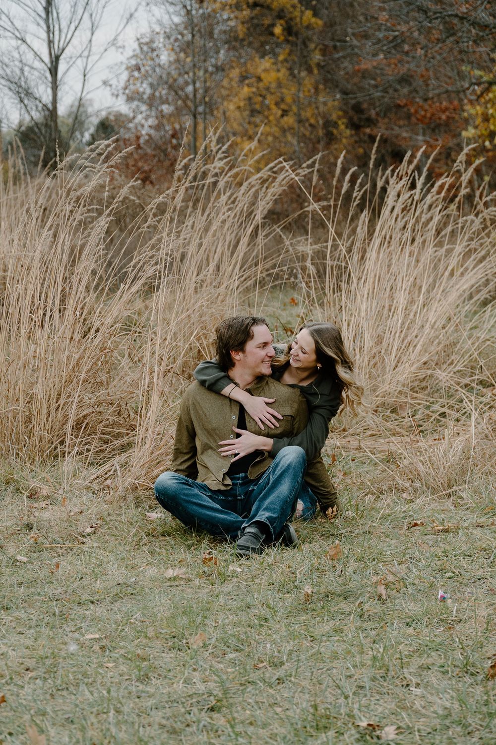 Intimate couple lying together in golden grass field during autumn season with trees in background.