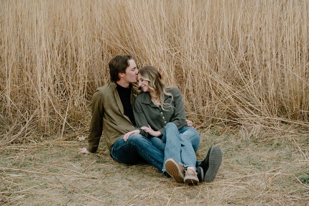 A couple sits together in tall grass during a romantic outdoor photoshoot at sunset.