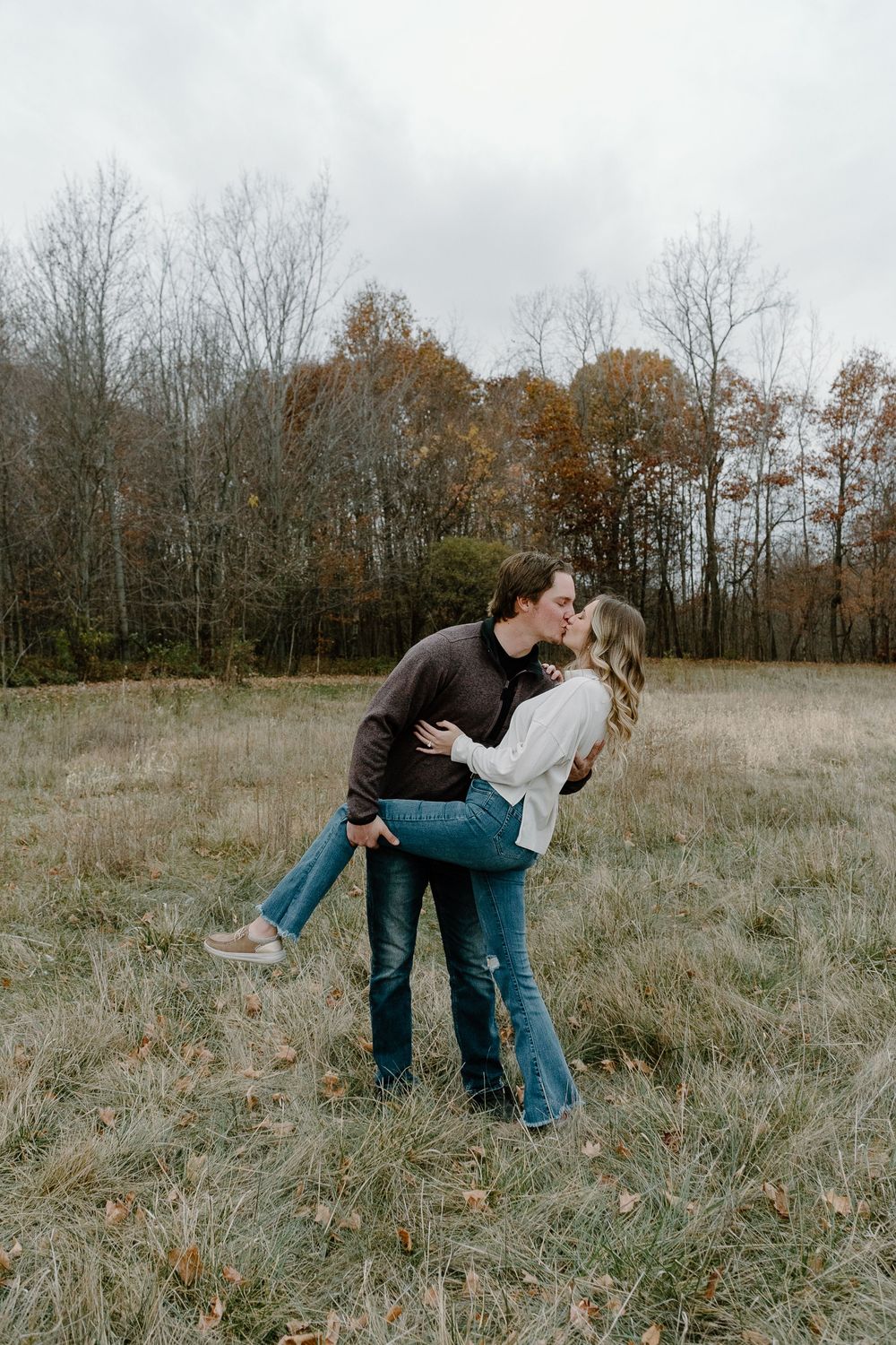 A couple shares romantic moments in a series of dips across an autumn field with bare trees in the background.