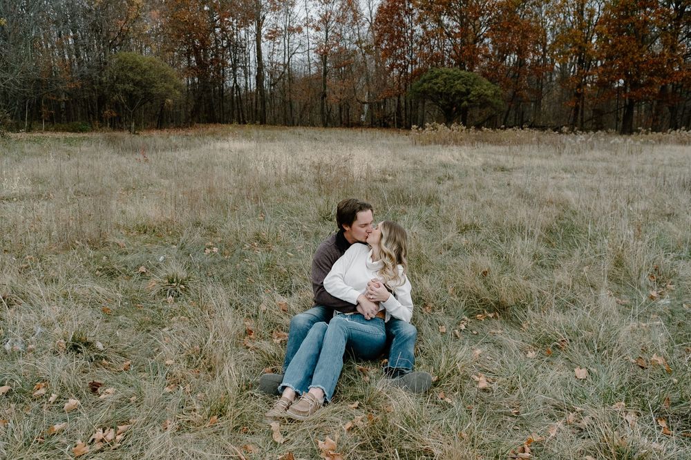 A couple sits together in a serene autumn field surrounded by dried grass and bare trees in the distance.