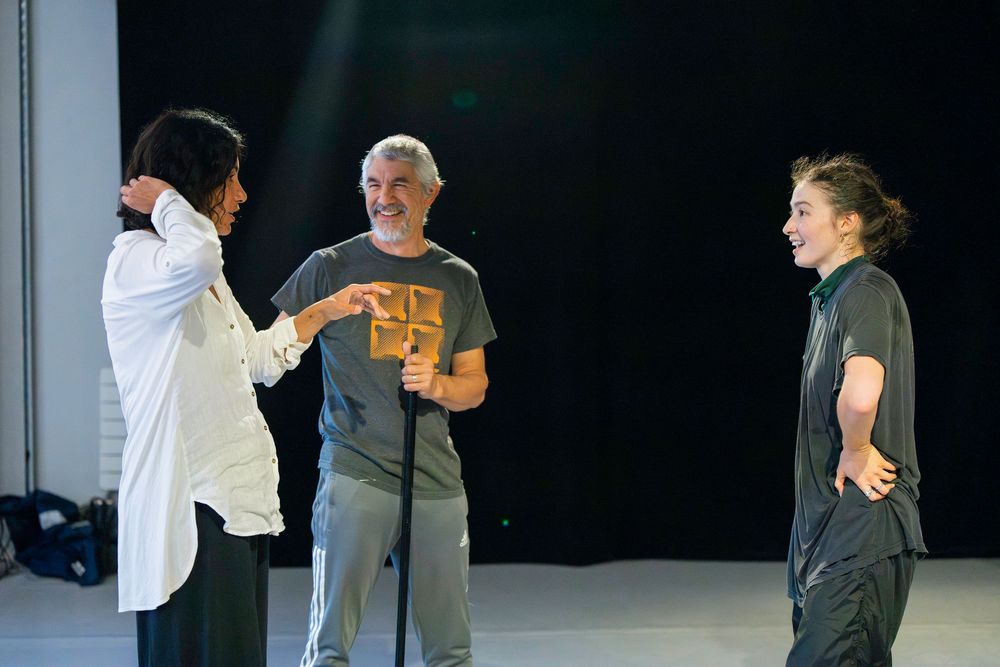 A series of dance performers in gray and white outfits practicing choreography on a dark stage.
