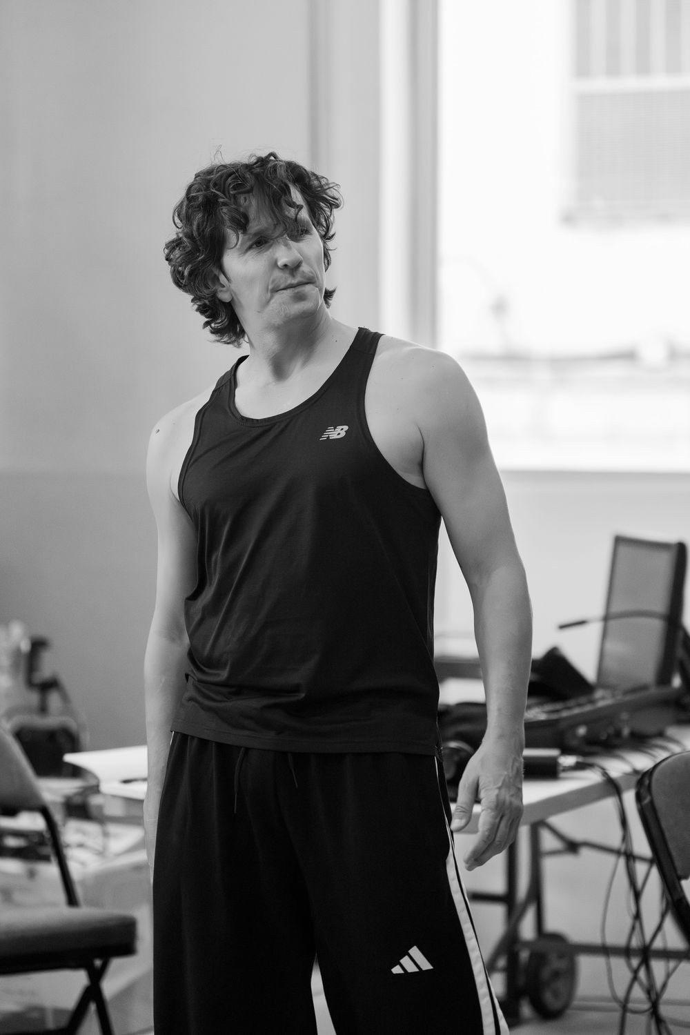 Performer in black tank top stands in rehearsal studio with natural lighting streaming through windows.