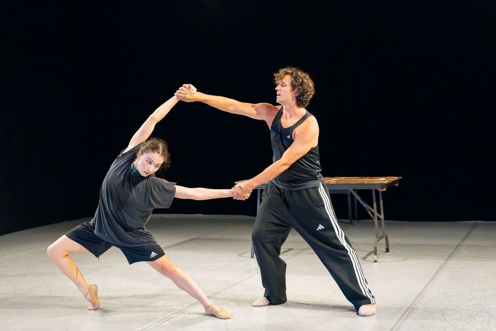 Two dancers perform a contemporary duet with flowing movements on a dark stage.