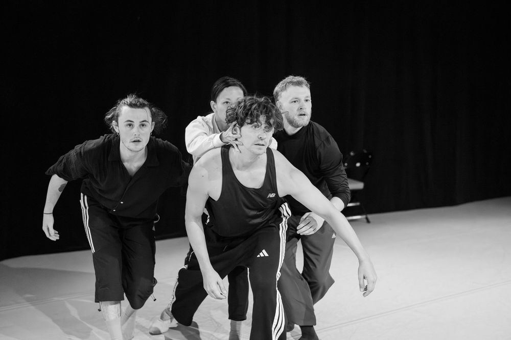 Three dancers in black attire perform a synchronized movement sequence on a white stage floor.