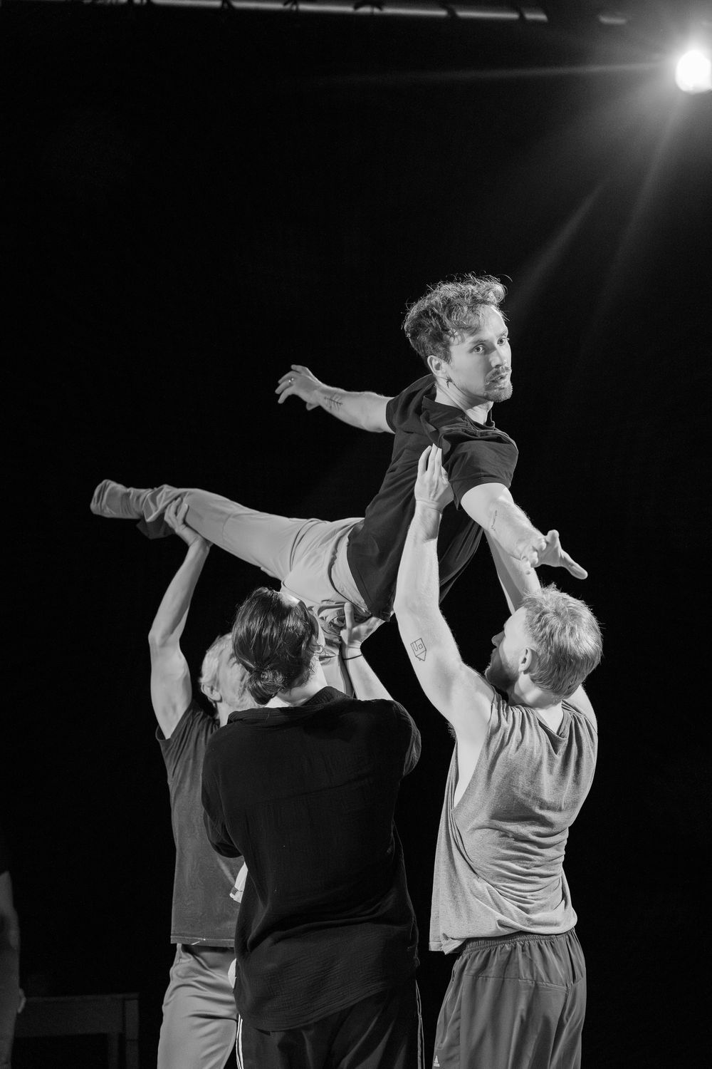 Black and white photo of dancers performing a lift during rehearsal on a dark stage with dramatic lighting.