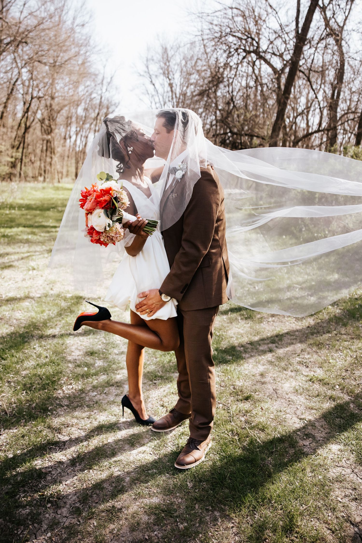 Interracial couple kissing under flowing veil in sunny park, bride holding colorful bouquet.