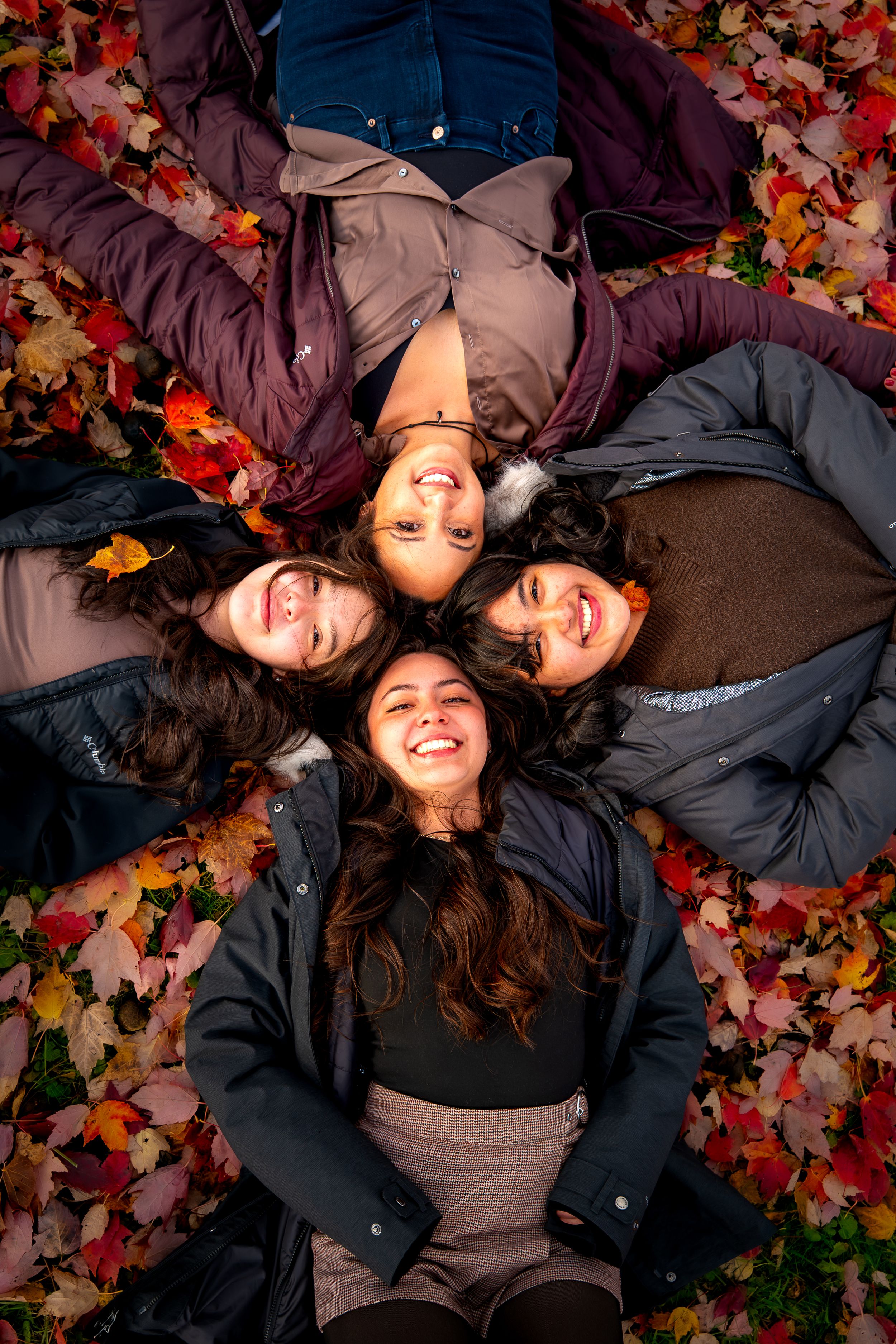 Four friends lying on colorful autumn leaves in a circle formation smiling at the camera.